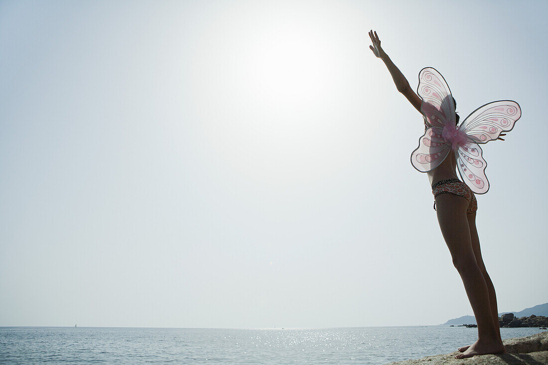 Girl in bikini wearing butterfly costume, standing on edge of cliff with arms raised