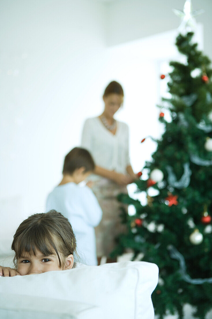 Girl looking over sofa at camera, boy and mother decorating Christmas tree in background
