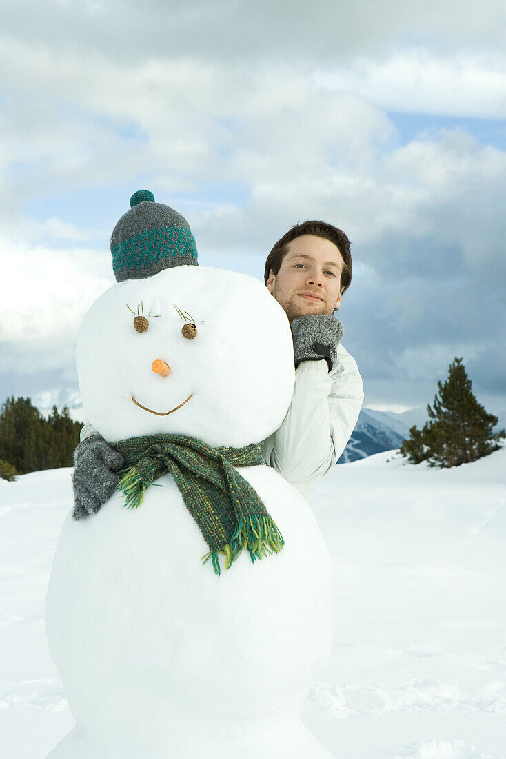 Young man embracing snowman, hand under chin, portrait