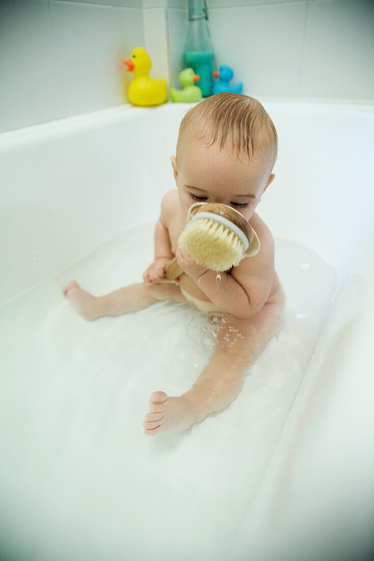 Baby taking bath, holding bath brush against face, full length