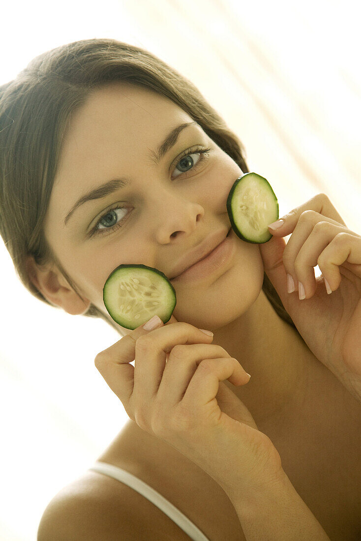 Young woman holding cucumber slices against face, smiling at camera