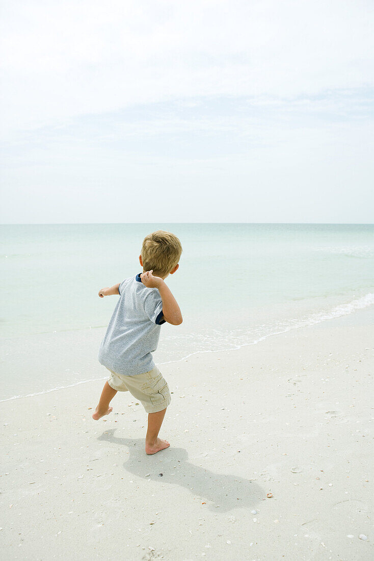 Boy on beach throwing unseen object toward ocean, rear view, full length