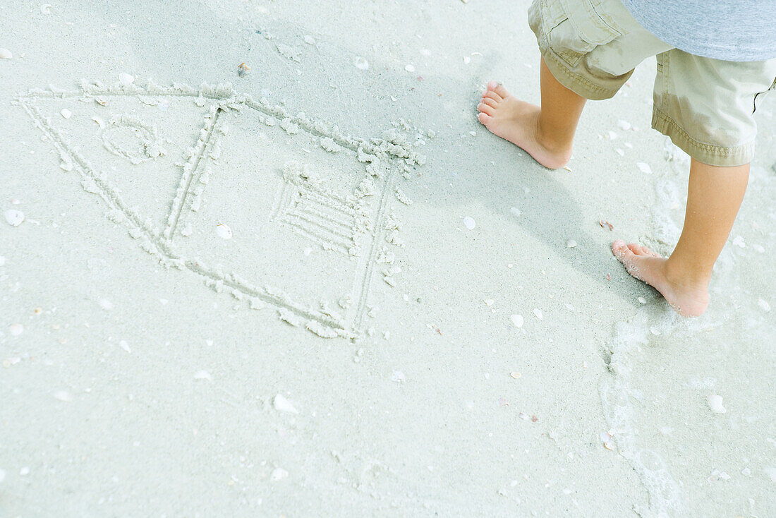 Child at the beach, standing next to drawing of house in sand, cropped view