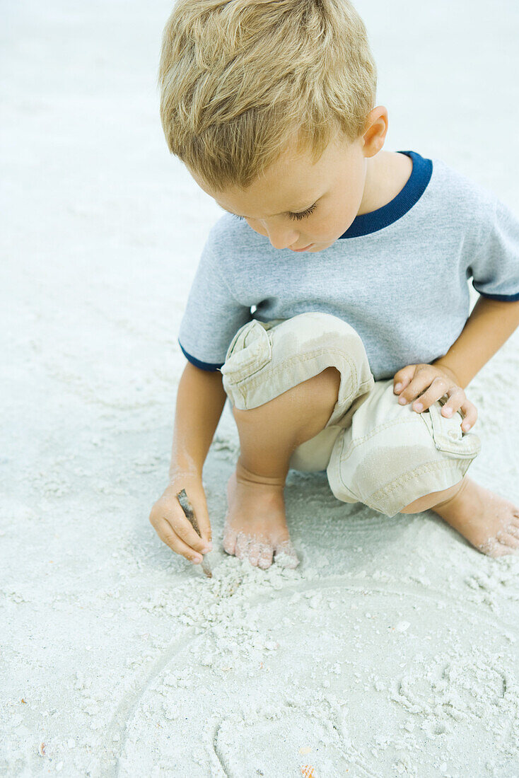 Little boy crouching at the beach, drawing in sand with stick