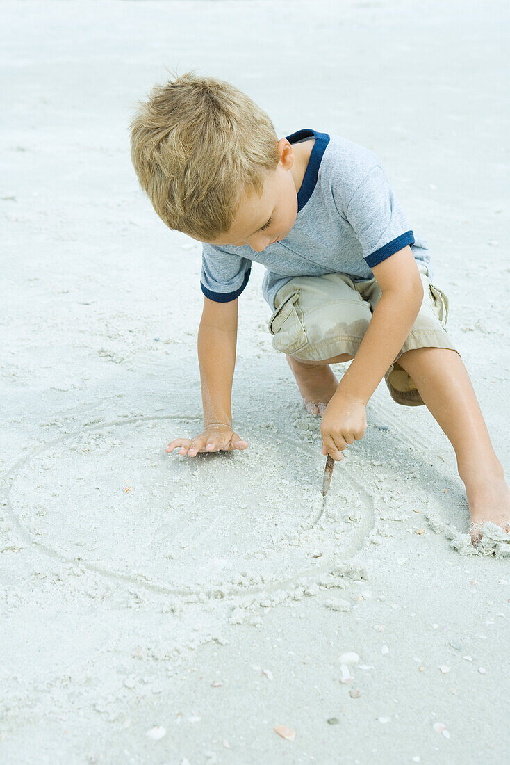 Little boy crouching at the beach, drawing in sand with stick