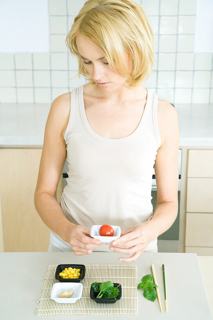 Young woman standing in kitchen, holding cherry tomato in small dish, looking down