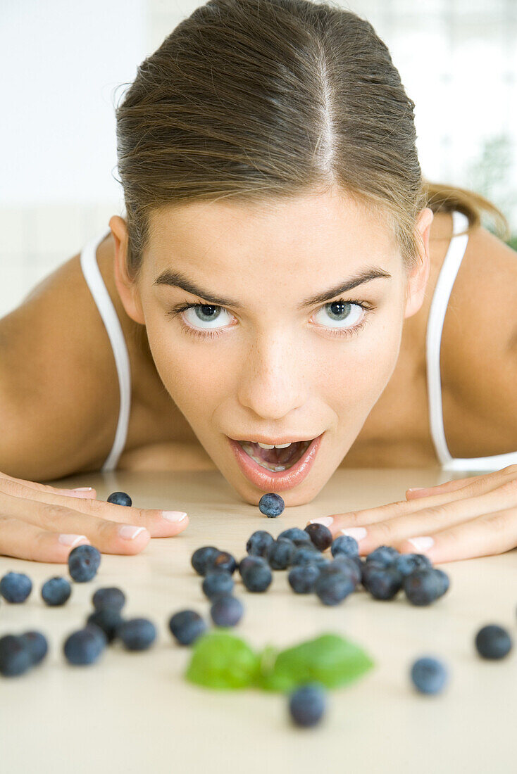 Fresh blueberries scattered across counter, young woman opening mouth to eat one