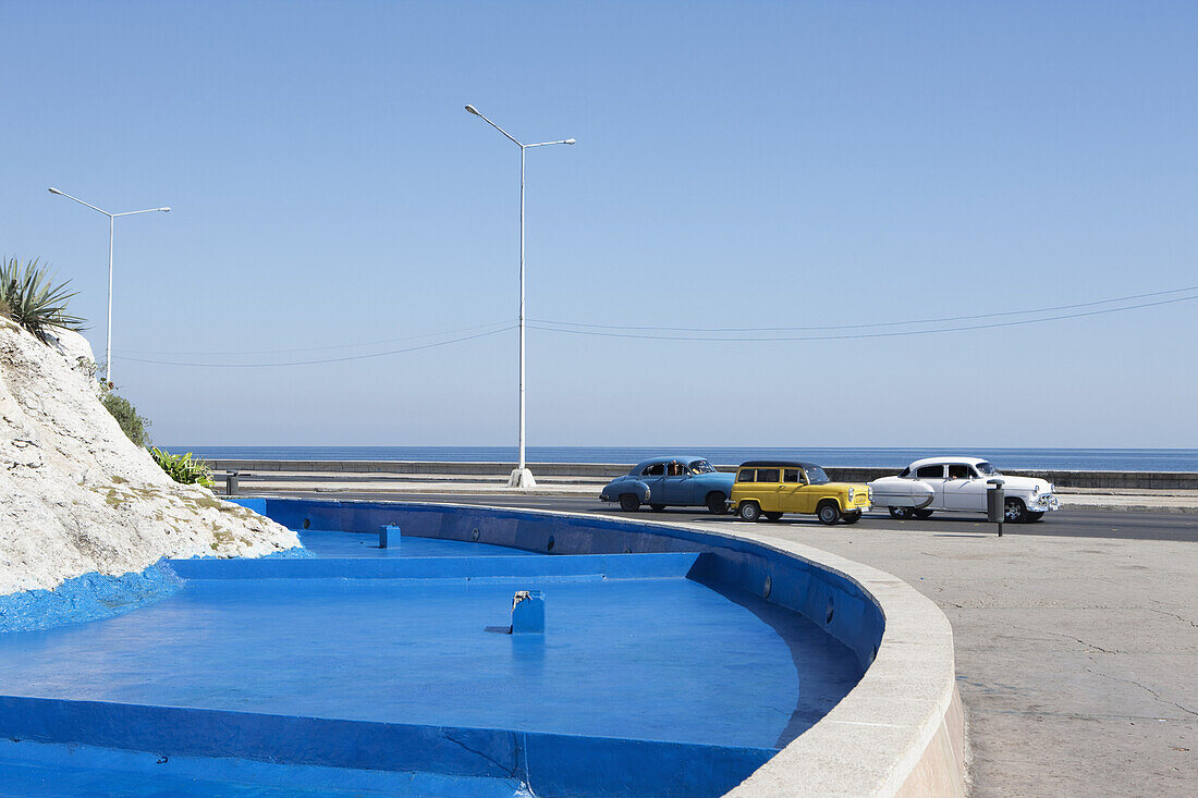 Classic cars moving by blue fountain against clear sky