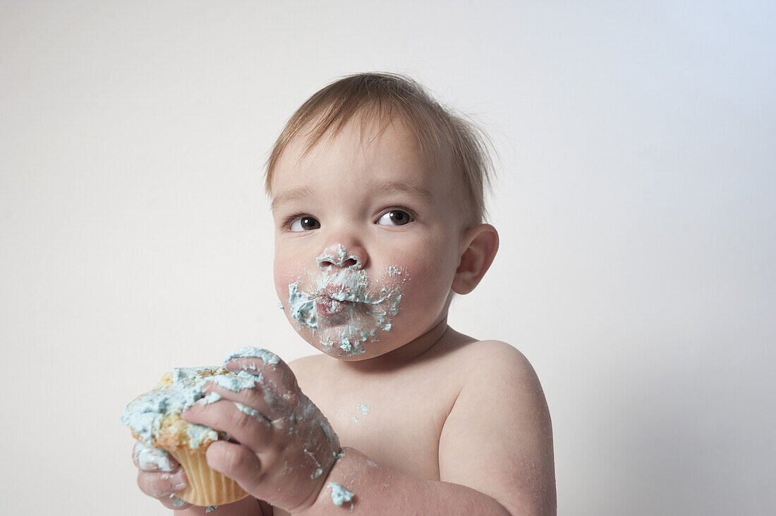 Cute baby boy with messy mouth looking away while holding cupcake against white background