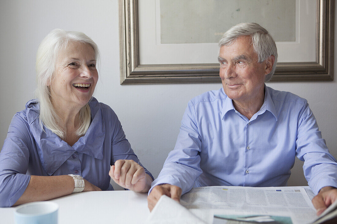 Happy senior couple with newspaper sitting at table in house