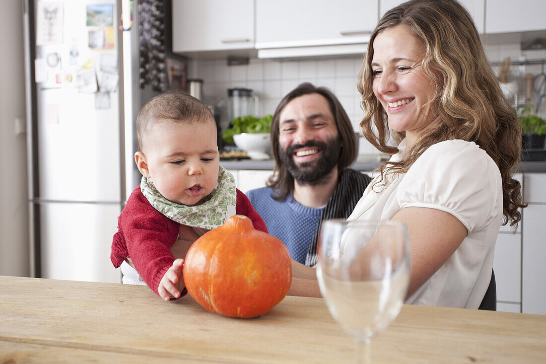 Happy parents looking at baby girl playing with pumpkin in kitchen
