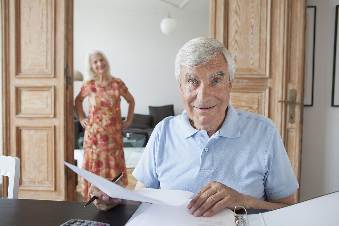 Portrait of surprised senior man checking financial documents at home with woman in background