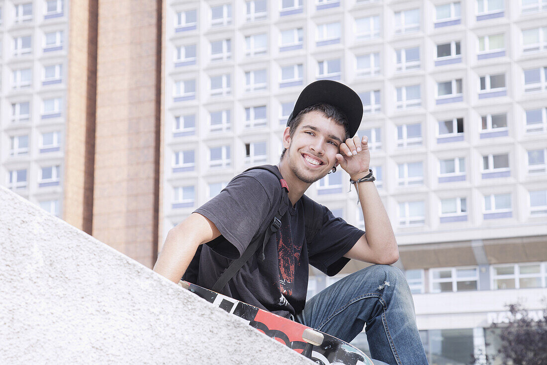 Happy young man with skateboard sitting against building