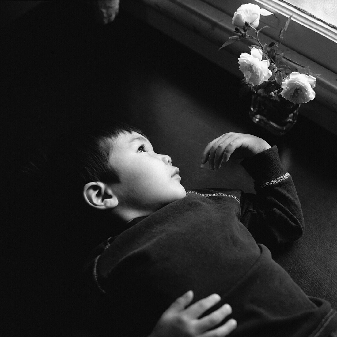 A young boy lying next to a vase of roses, staring out window