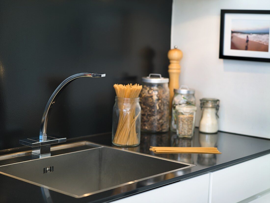 A stainless steel sink with storage jars against a black wall