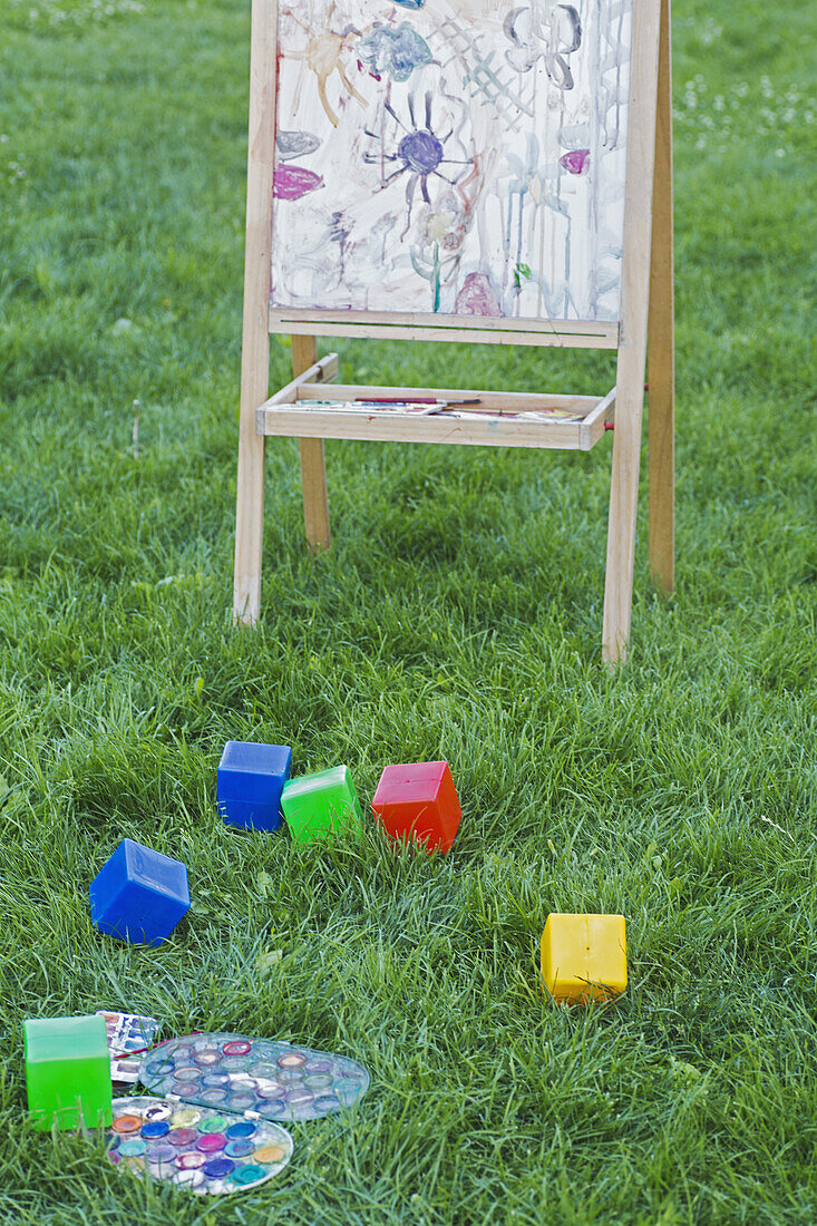 An easel, toy blocks and watercolor paints in the grass of a backyard