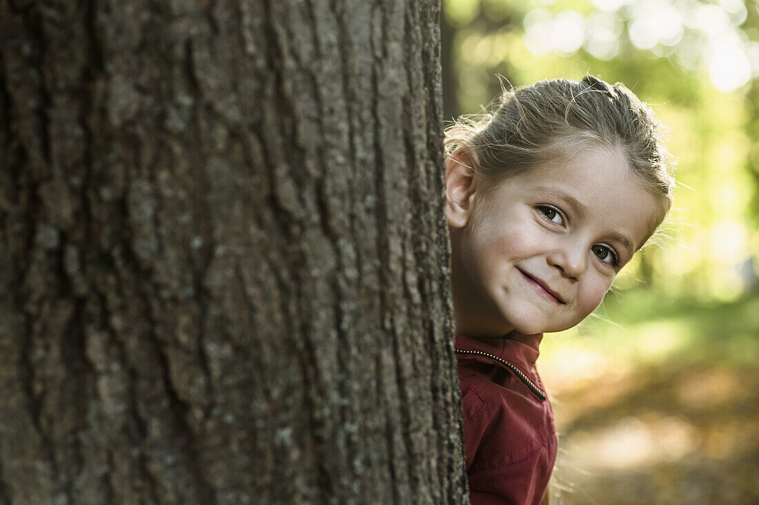 A young smiling girl peeking from behind a tree trunk