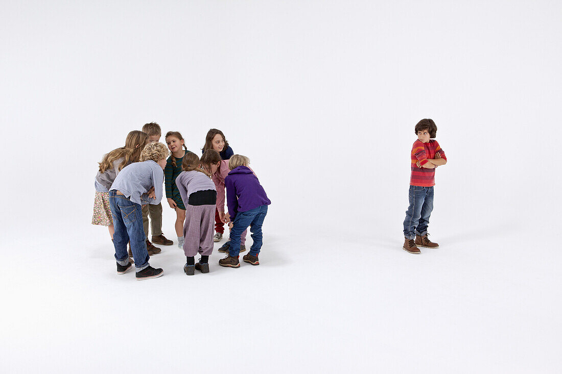 A boy standing alone, arms crossed, while a group of kids gossip about him