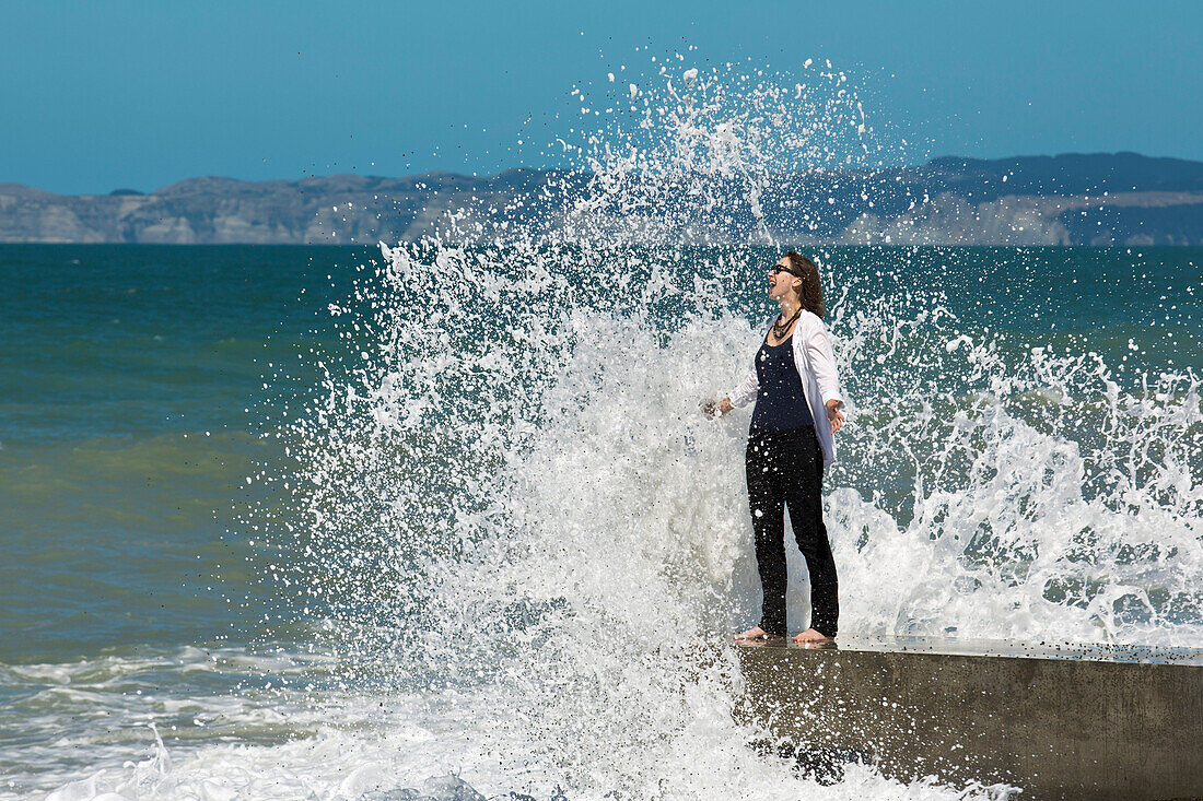 Happy woman standing by the sea and getting sprayed by a wave (MR), Napier, Hawke's Bay, North Island, New Zealand