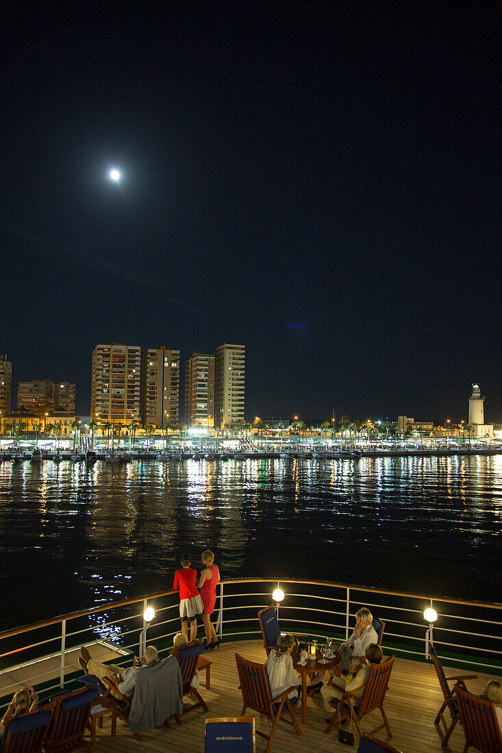 Passengers sitting outside bar Zum Alten Fritz aboard cruise ship MS Deutschland (Reederei Peter Deilmann) as the moon rises over apartment buildings at night, Malaga, Andalusia, Spain