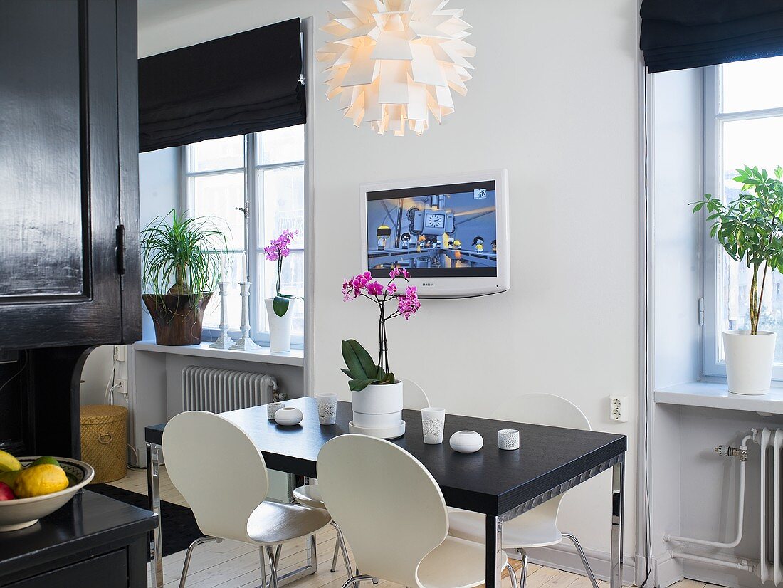 A black dining table with white Bauhaus chairs and a TV on the wall