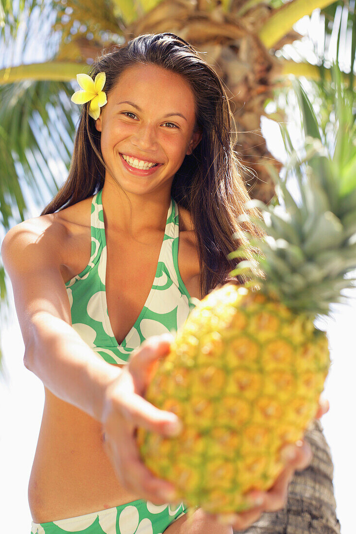 Teenage Girl Wearing A Bikini Holding Out A Pineapple, Flower In Her Hair, Palm Tree In Background.