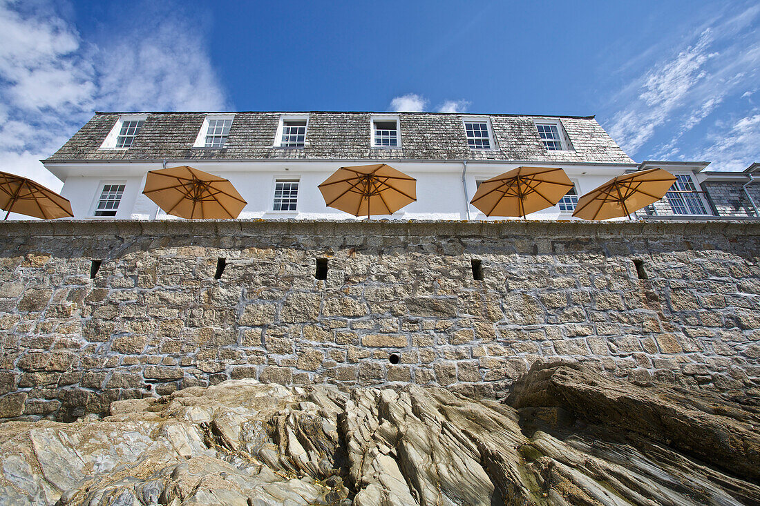 'Looking up a wall from the shore to a building and umbrellas on a patio on the seaside; St. Mawes, Cornwall, England'