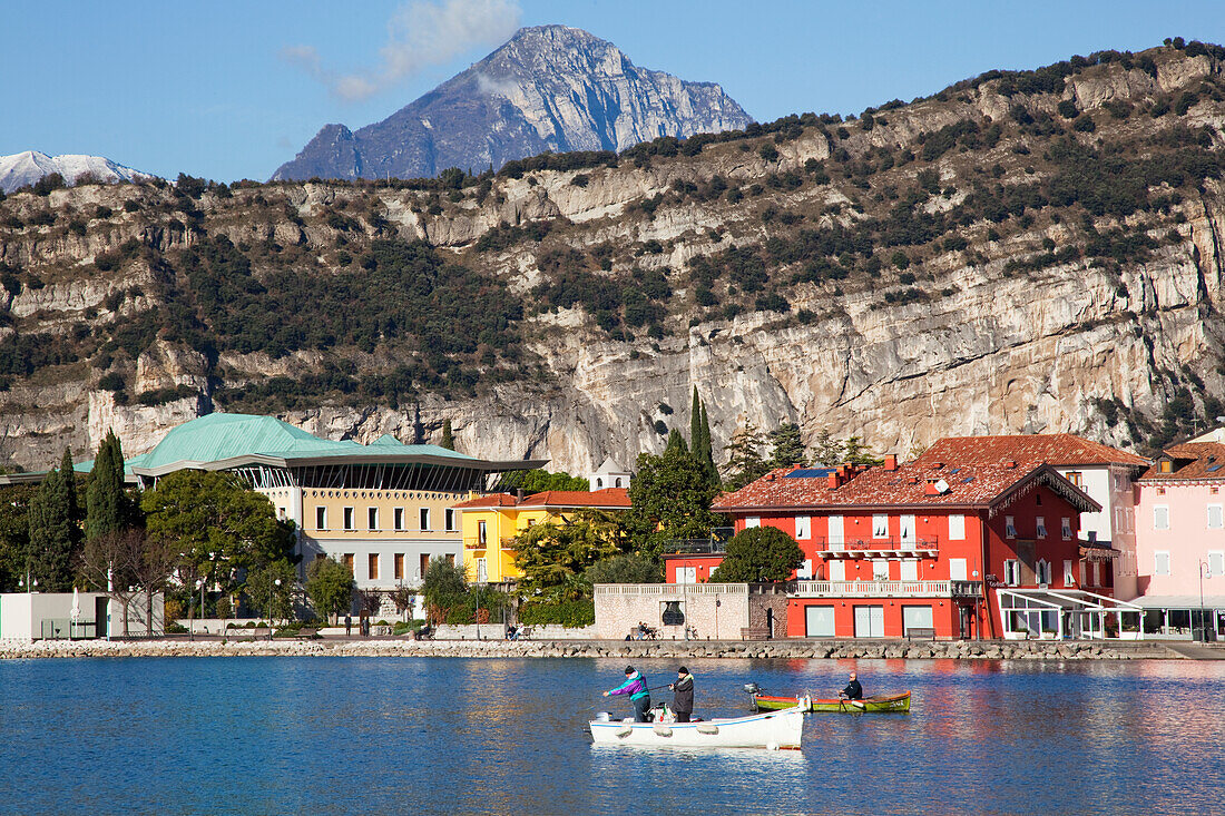 'Cliffs and buildings along the shoreline of Lake Garda with men fishing in the harbour; Torbole, Trentino, Italy'