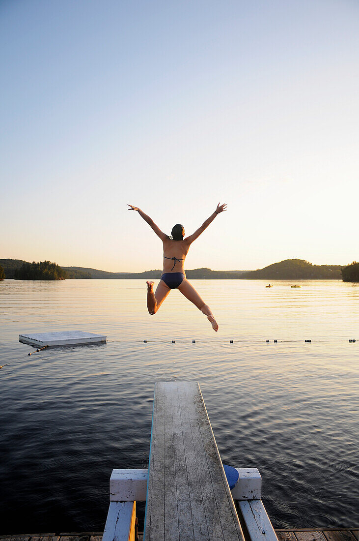 'A swimmer jumps off a diving board as the sun sets over a lake in Muskoka; Huntsville, Ontario, Canada'