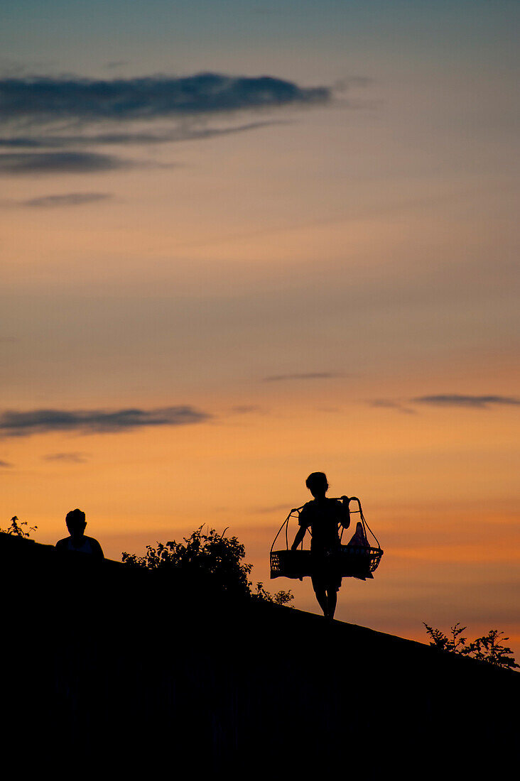 A boy walking over a bridge in silhouette at sunset carrying baskets over his shoulders.  Laos