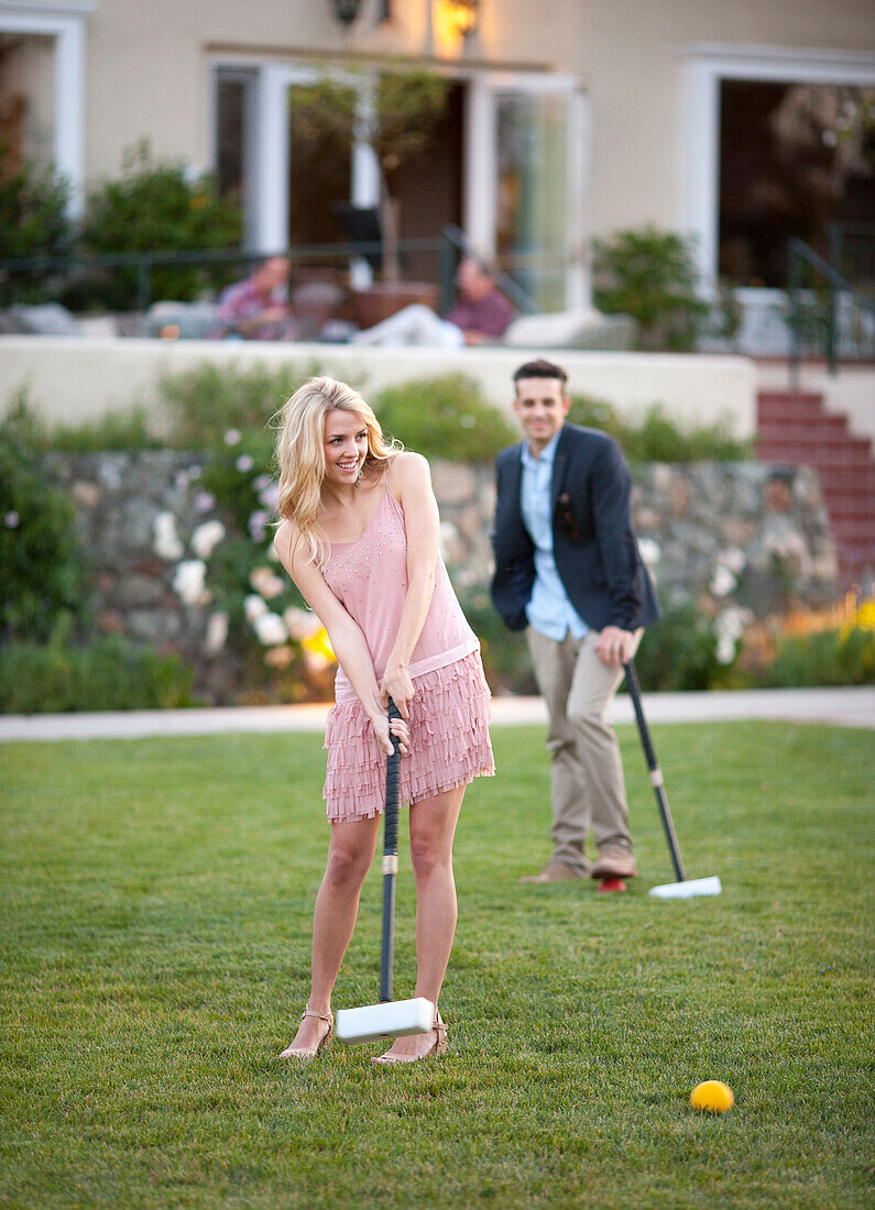 A young beautiful lady hit a croquet ball with a mallet, her male partner stands at a distance with a mallet in his hand observes her.