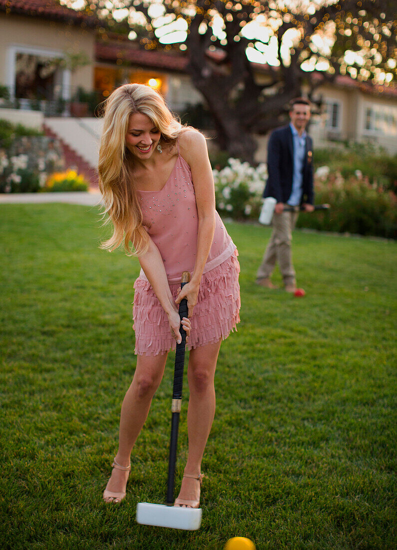 A beautiful young lady plays croquet, her male partner watches from behind.