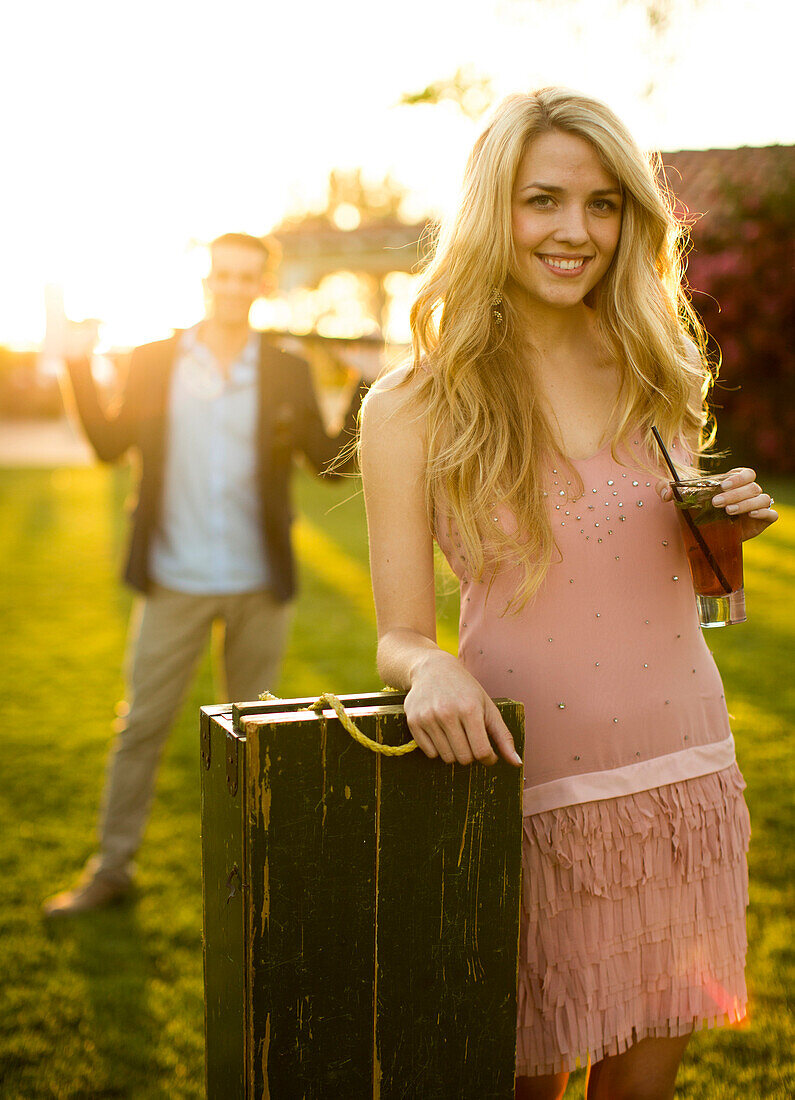 A beautiful lady with a drink glass on her left hand stands against a croquet box with her right elbow rests on it, her male partner stands behind her at a distance.