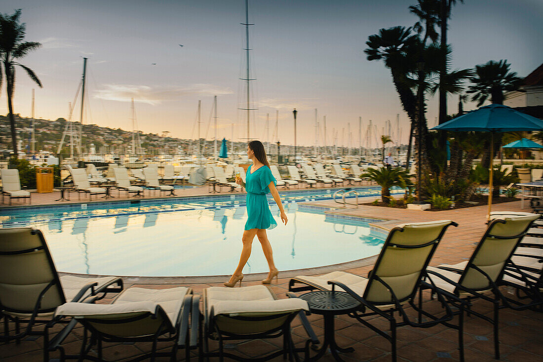 A young girl strolls by the swimming pool with her drink.