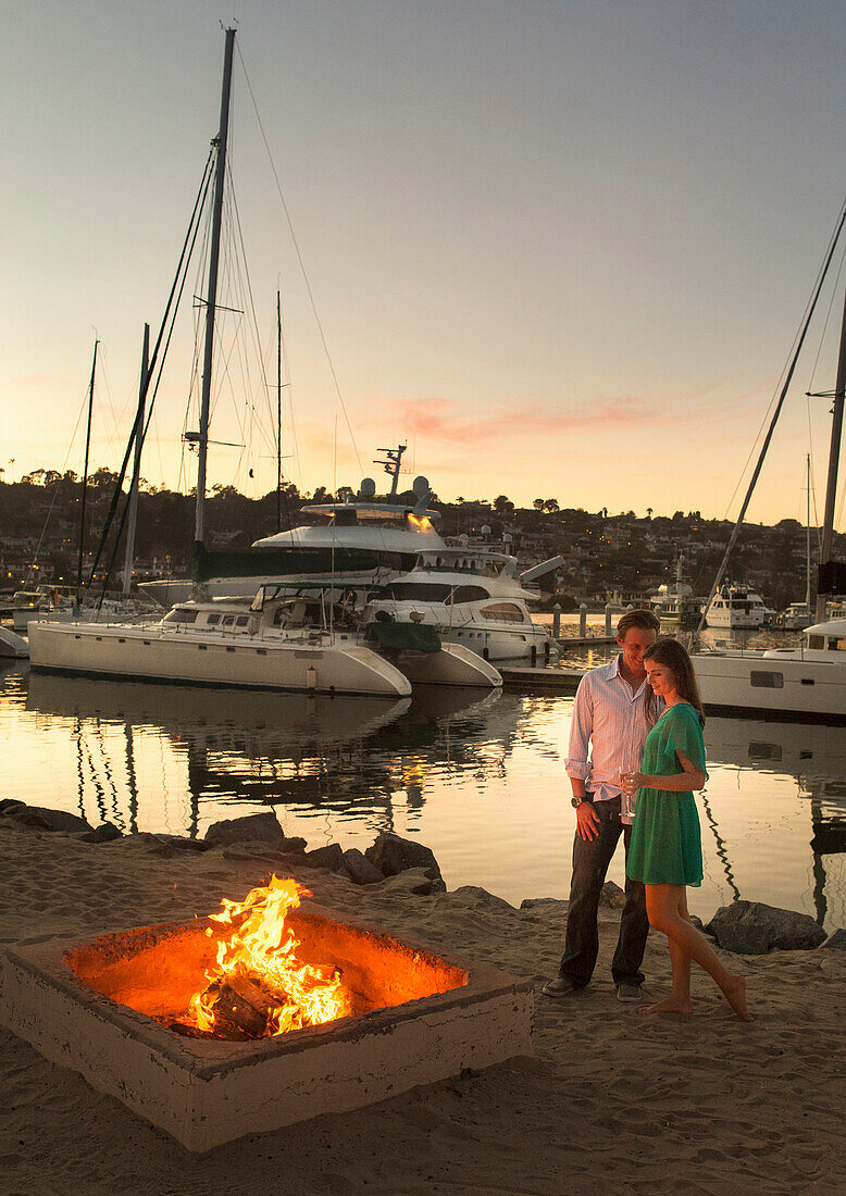 A young girl with a drink in her left hand stands intimately with her male partner next to a fireplace by the marina.