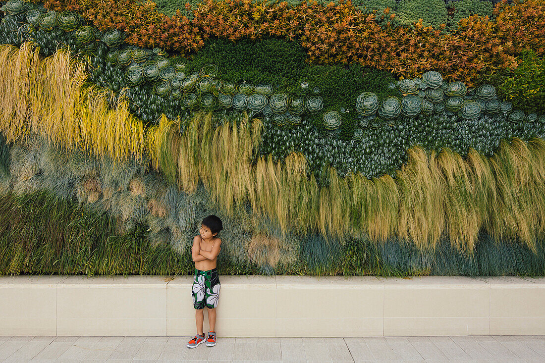 A little boy stands crossed arms by flower wall.