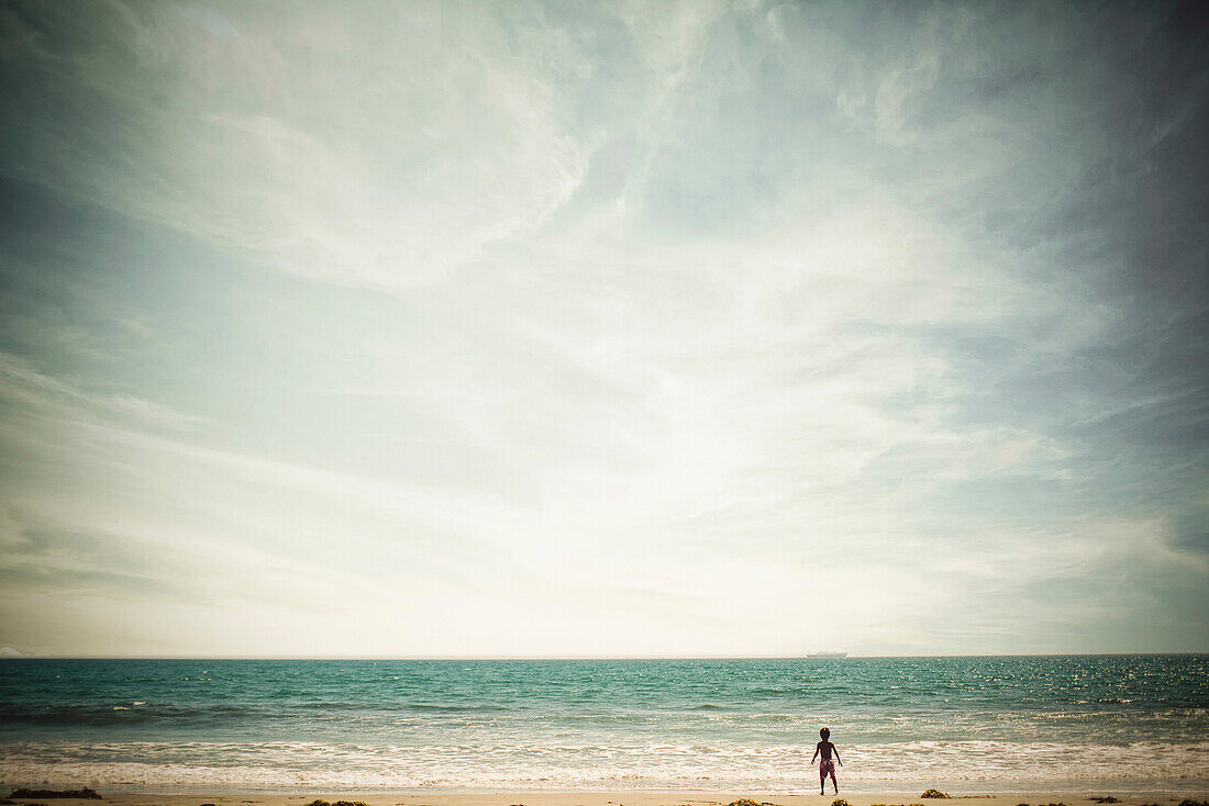 A young boy standing on a California beach looking out over the ocean.