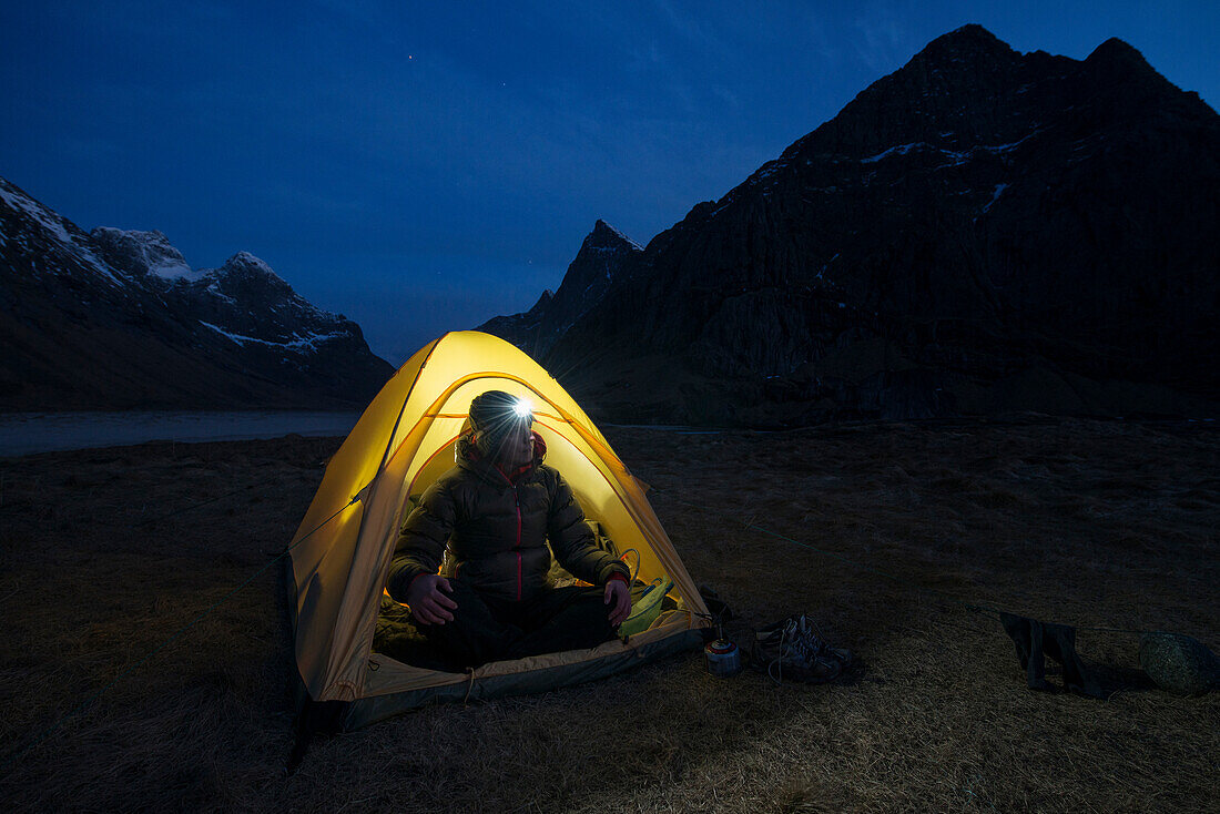 Female hiker sits in tent illuminated at night while wild camping at scenic Horseid beach, Moskenes??y, Lofoten Islands, Norway