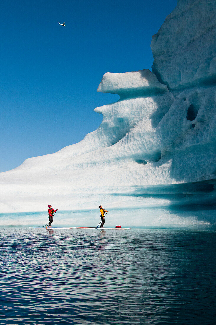 As a small plane flies overhead, two adults on stand up paddle boards (SUP) view an iceberg on Bear Lake in Kenai Fjords National Park, Alaska.