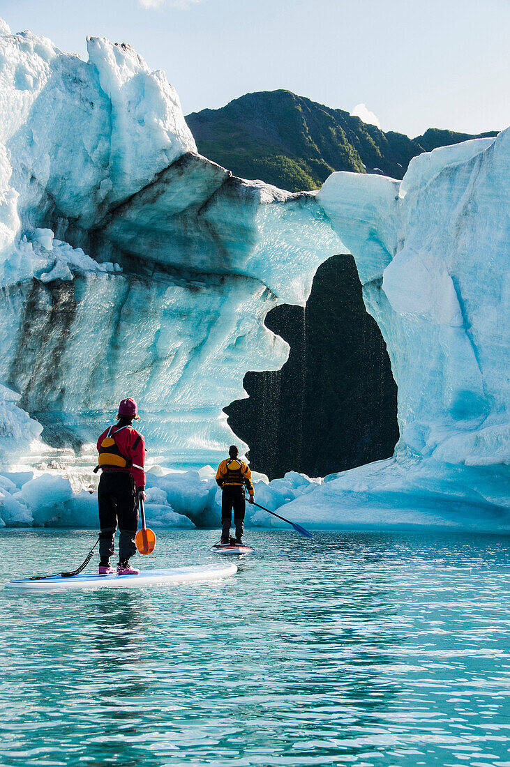 Two adults on stand up paddle boards (SUP) observe hole melted in iceberg on Bear Lake in Kenai Fjords National Park, Alaska.