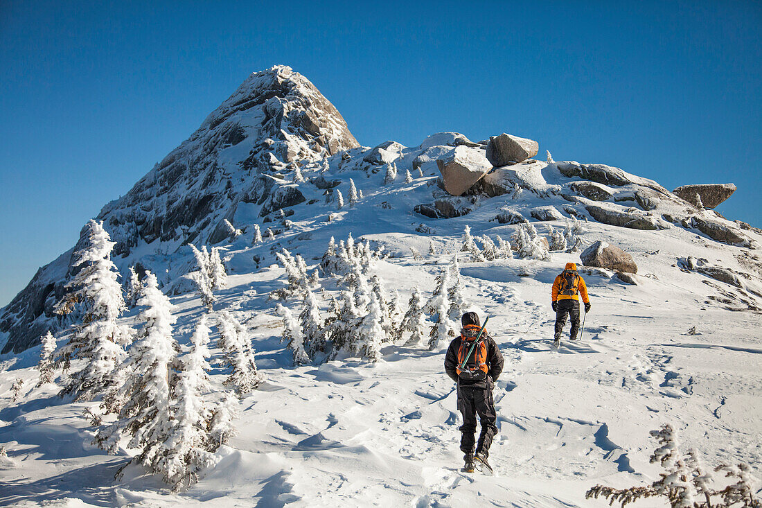 Two climbers wearing backpacks walk a wide snow-covered ridge toward Needle Peak in the Coquihalla Recreation Area of British Columbia, Canada.