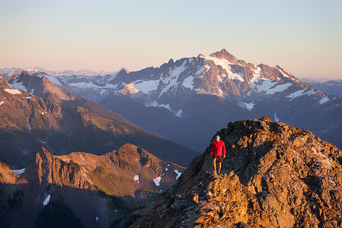 A hiker walks a rocky ridge with Mount Shuksan in the background.