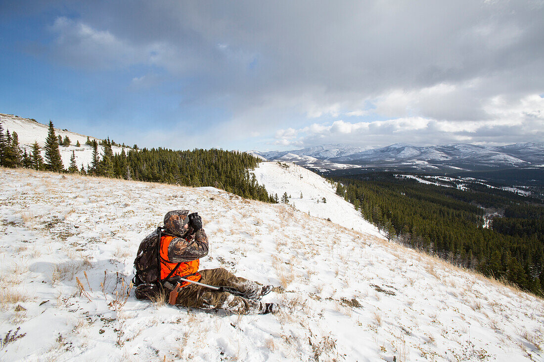 A male hunter looks through binoculars in the snow.