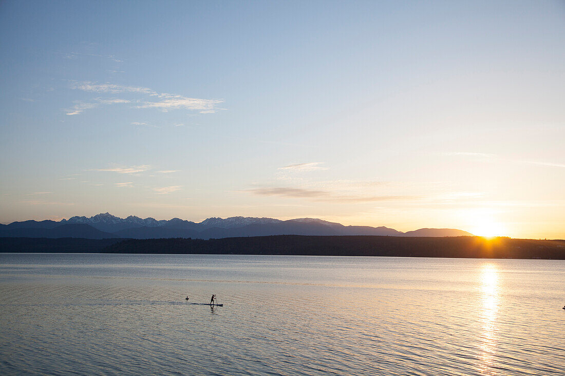 A fit male paddles his stand up paddle board (SUP) along the Hood Canal in the Puget Sound with the Olympic Mountains behind him near Poulsbo, Washington at sunset.