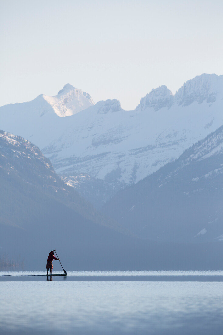 A man stand up paddle boards (SUP) on a calm Lake McDonald at sunrise in Glacier National Park near West Glacier, Montana.