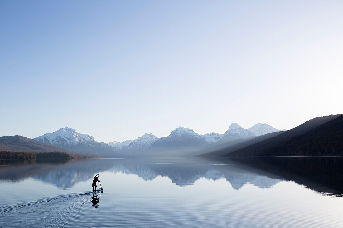 A man stand up paddle boards (SUP) on a calm Lake McDonald at sunrise in Glacier National Park near West Glacier, Montana.