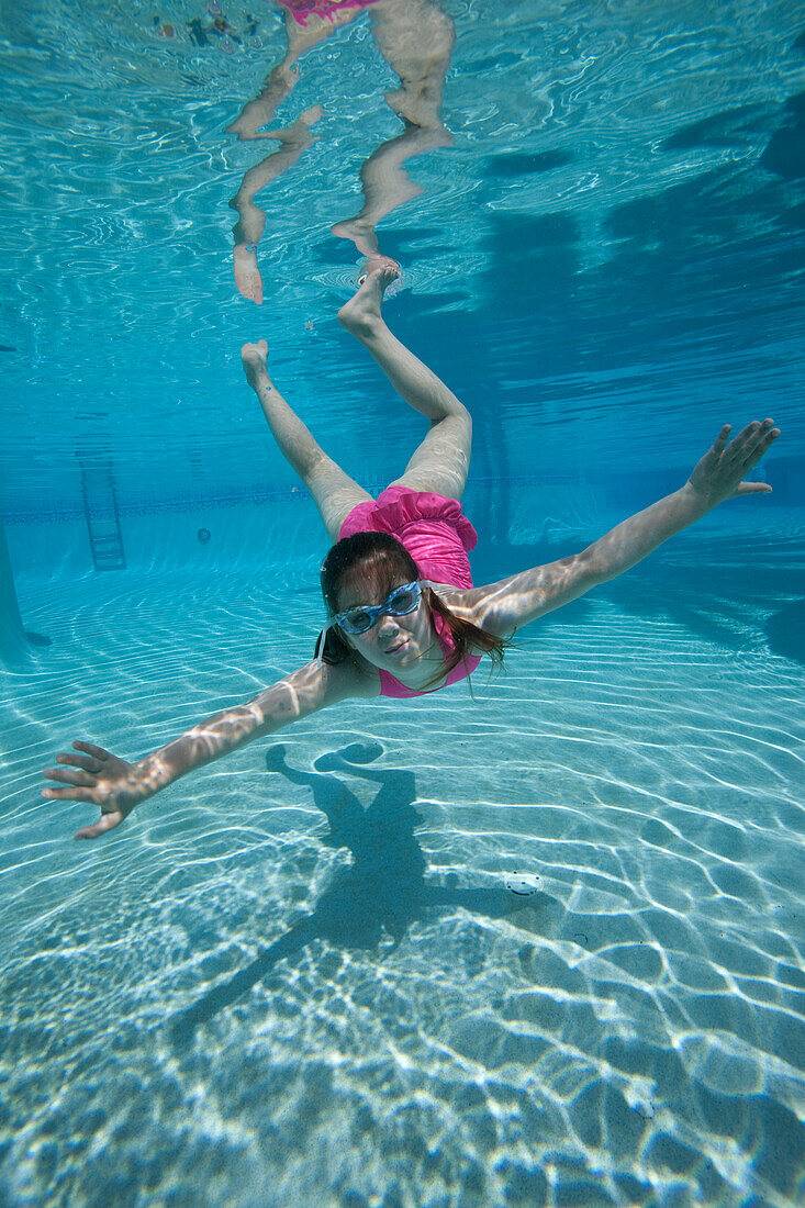 a girl swimming toward camera underwater in pool wearing a pink swimsuit and goggles