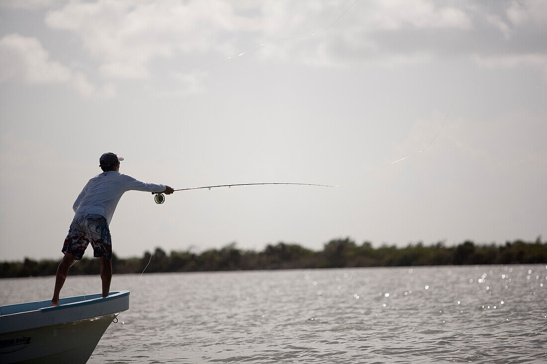 a fly fisherman casts his rod while standing on the bow of a boat with mangroves in the background