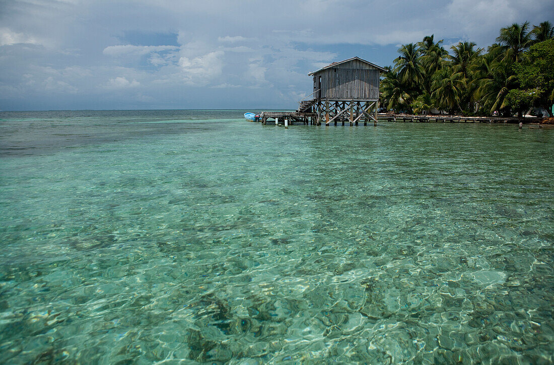 a wooden fishing hut sits above turquoise waters with palm trees growing on island