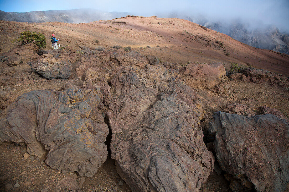 A woman in her thirties carrying an infant hikes in the high-elevation volcanic Haleakala crater, with ropy pahoehoe lava in the foreground.